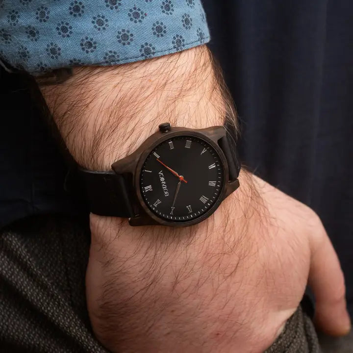 Black wristwatch with a detailed face on a person's wrist, wearing a blue shirt.