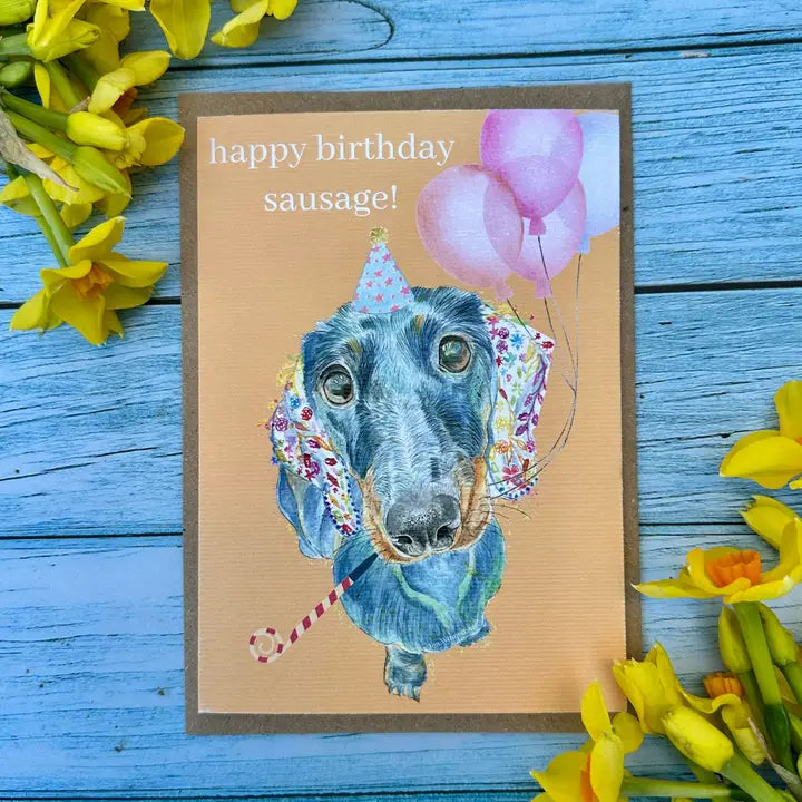 Birthday card with a dog illustration, balloons, and 'happy birthday sausage!' text on a wooden surface with yellow flowers.
