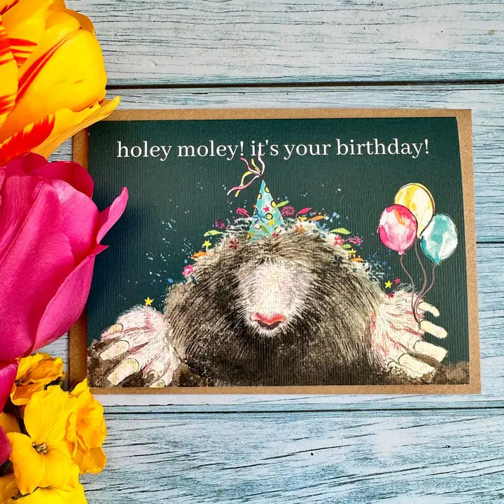 Birthday card with a moles illustration and colorful text, surrounded by flowers on a wooden surface.