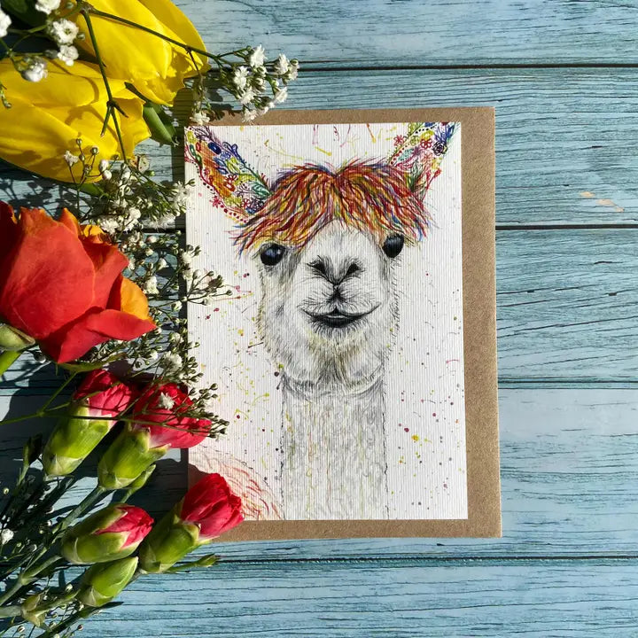 Greeting card with a llama illustration on a wooden surface with flowers.