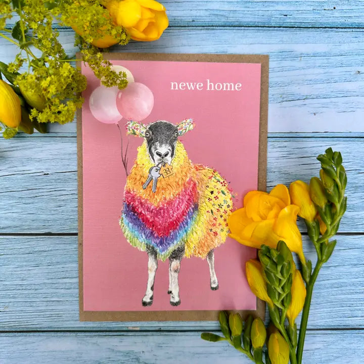 Greeting card with a colorful sheep illustration and 'new home' text, surrounded by yellow flowers on a blue wooden surface.