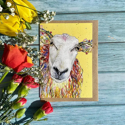 Card with a colorful sheep illustration on a yellow background, surrounded by flowers on a wooden surface. card