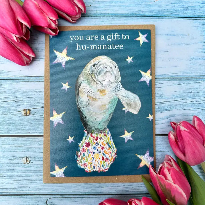 Card with a manatee illustration and text surrounded by pink tulips on a wooden surface