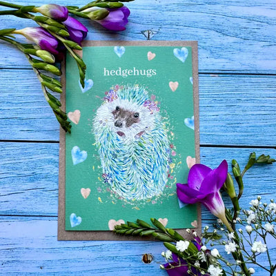Card with hedgehog illustration and 'hedgehugs' text on a blue wooden background with flowers.